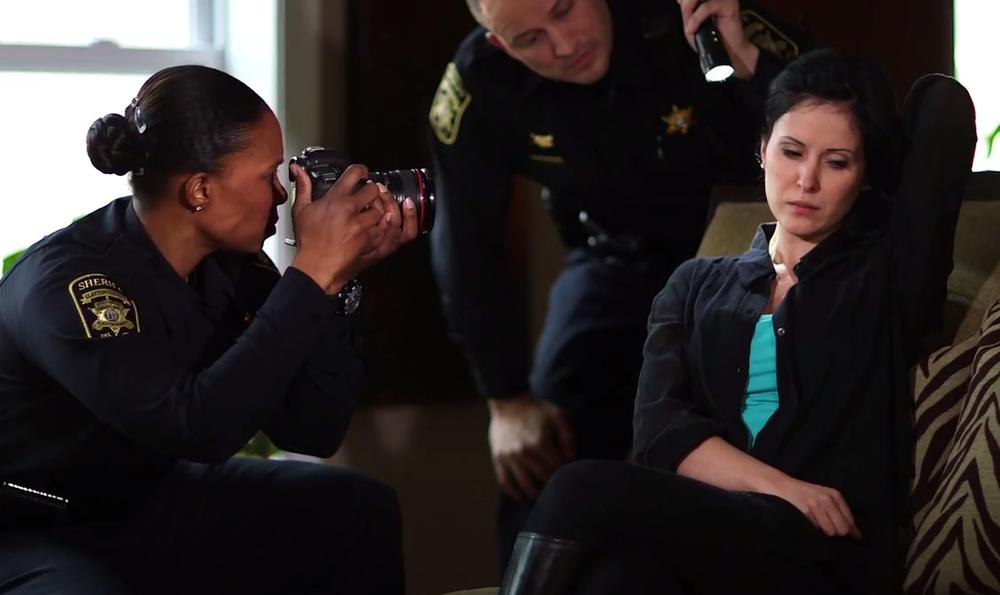 an image of a woman holding her hair back, a police officer shines a flashlight on her neck while another takes a photo. 